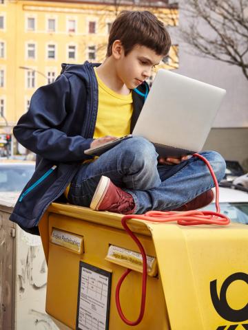 Junge sitzt mit Laptop und Kabelschnur auf einem Briefkasten