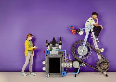 Ein Mädchen tippt auf dem Smartphone das an einer Riesenmaschine angeschlosse ist, auf der ein Junge sitzt